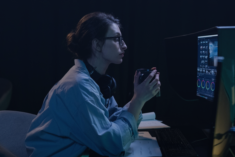 A woman in a dimly lit room working intently on a computer while holding a mug.