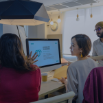 A diverse group of professionals working together in a contemporary office setting using laptops and a monitor.